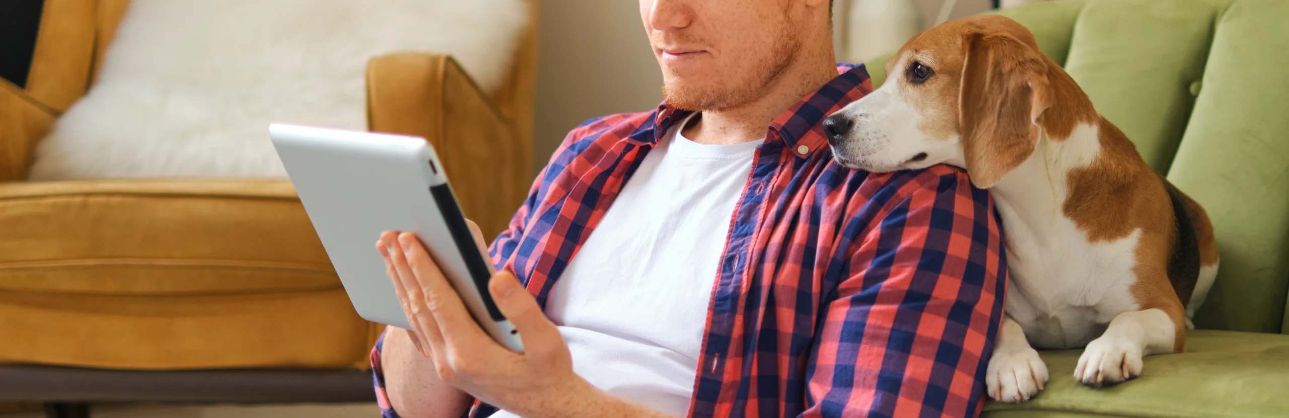 Man sitting on a couch using a tablet while a dog rests beside him and looks at the screen.